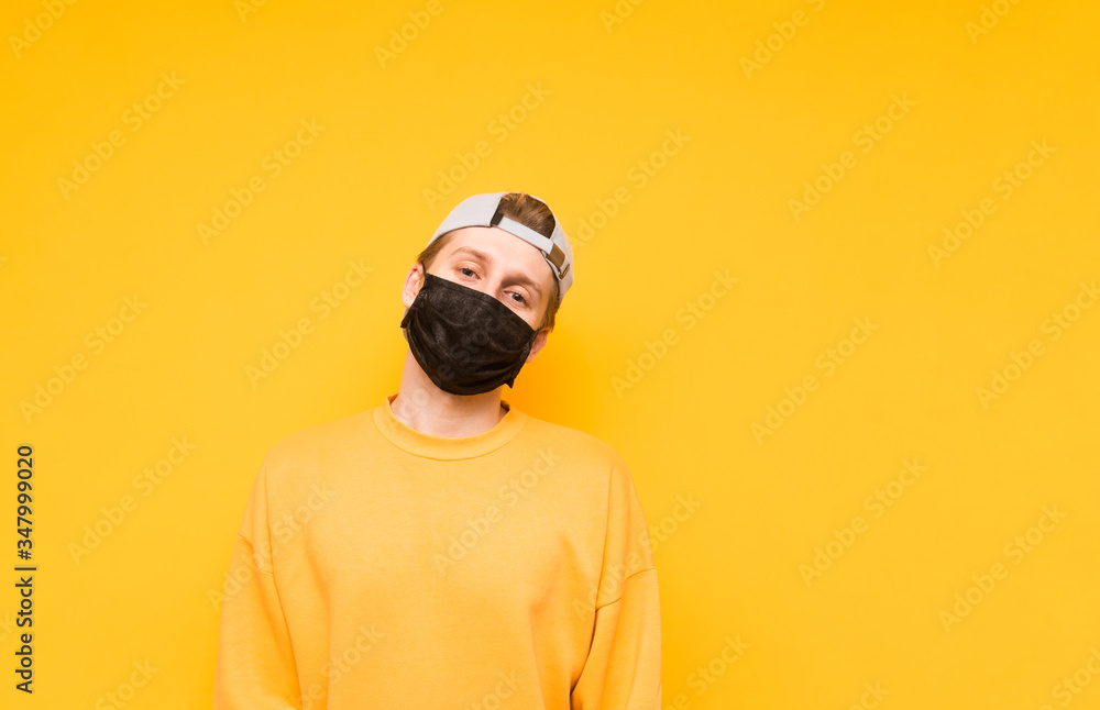 The guy in a cap and a black medical mask stands on a yellow background and poses for the camera with a serious face. Portrait of a young man in a protective gauze mask. Isolated. Coronavirus pandemic The guy in a cap and a black medical mask stands on a yellow background and poses for the camera with a serious face. Portrait of a young man in a protective gauze mask. Isolated. Coronavirus pandemic