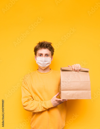 Happy young man in a protective mask stands on a yellow background and holds in his hand a paper bag with food from the delivery. Coronavirus pandemic. Quarantine. covid-19. Vertical