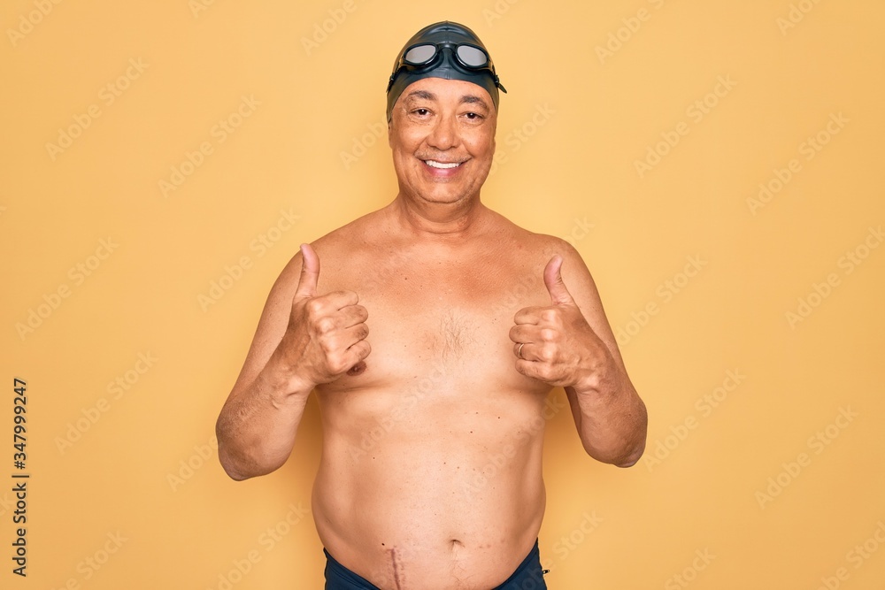 Middle age senior grey-haired swimmer man wearing swimsuit, cap and goggles success sign doing positive gesture with hand, thumbs up smiling and happy. Cheerful expression and winner gesture.