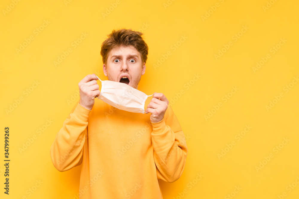 Frightened young man with a shocked face puts a medical mask on his face. Shocked guy puts on a gauze protective mask on a yellow background. Coronavirus pandemic. Quarantine. covid-19. Frightened young man with a shocked face puts a medical mask on his face. Shocked guy puts on a gauze protective mask on a yellow background. Coronavirus pandemic. Quarantine. covid-19.