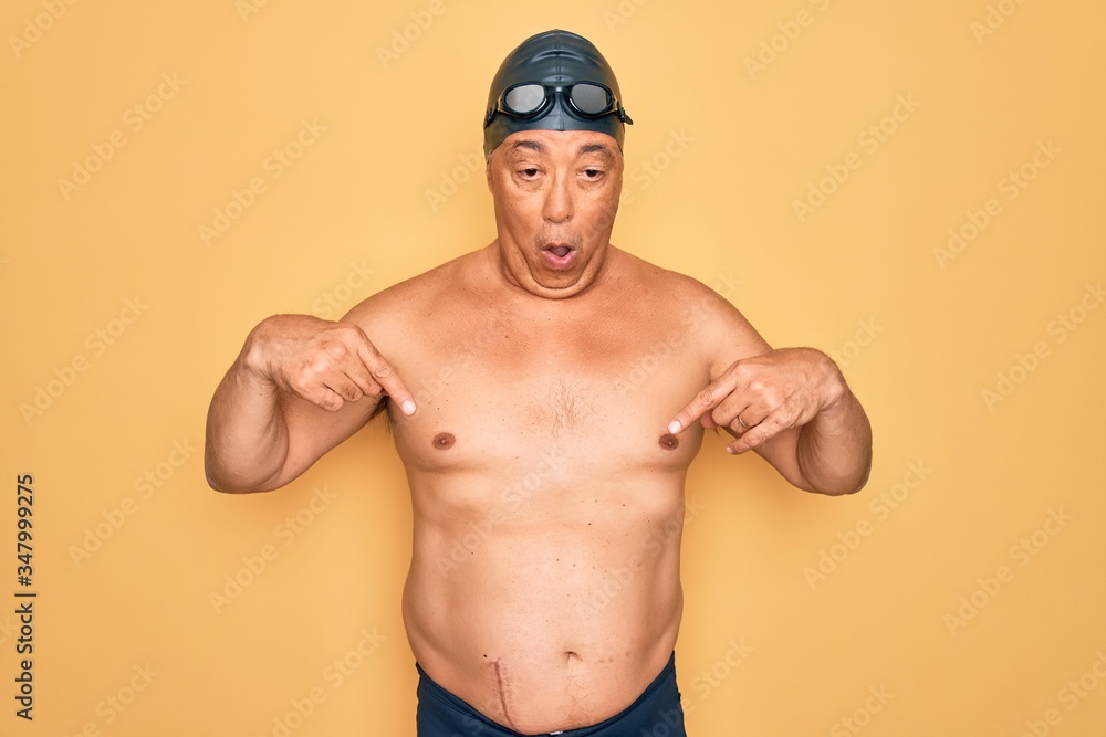 Middle age senior grey-haired swimmer man wearing swimsuit, cap and goggles Pointing down with fingers showing advertisement, surprised face and open mouth