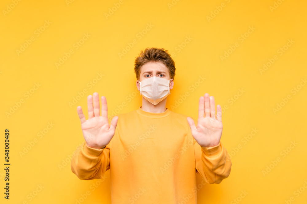 Young man in a medical mask stands on a yellow background, looks at the camera and shows his palms to the camera. The guy in a protective mask shows his hands with his palms. Quarantine. covid-19. Young man in a medical mask stands on a yellow background, looks at the camera and shows his palms to the camera. The guy in a protective mask shows his hands with his palms. Quarantine. covid-19.