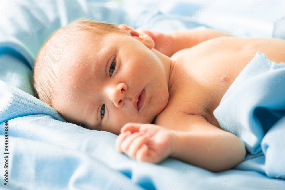 little caucasian newborn baby boy laying down on delicate blue blanket, falling asleep in safe, warm fabric. Adorable, sleepy new born baby opening his eyes.