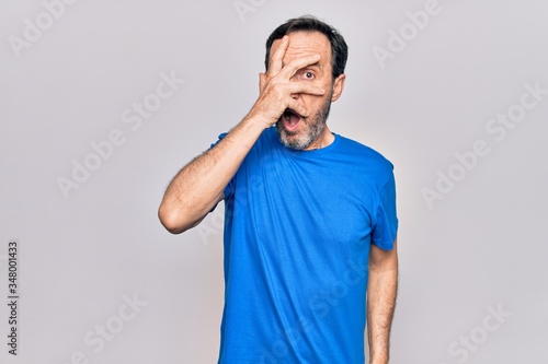 Middle age handsome man wearing casual t-shirt standing over isolated white background peeking in shock covering face and eyes with hand, looking through fingers afraid