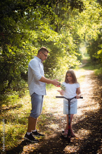 Happy friendly family of father and preteen girl riding scooter outdoors in the forest.