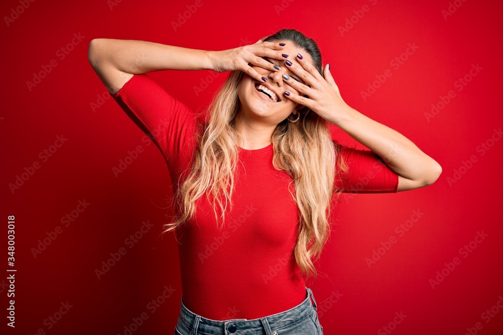 Young beautiful blonde woman wearing casual t-shirt standing over isolated red background covering eyes with hands smiling cheerful and funny. Blind concept. Young beautiful blonde woman wearing casual t-shirt standing over isolated red background covering eyes with hands smiling cheerful and funny. Blind concept.
