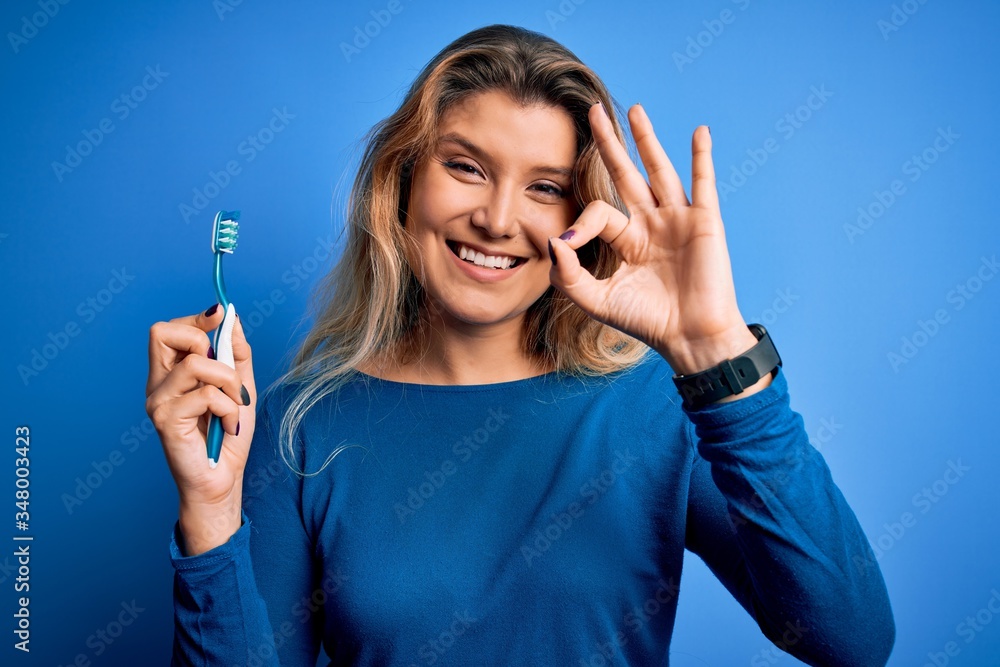 Young beautiful blonde woman using teeth brush over isolated blue background doing ok sign with fingers, excellent symbol Young beautiful blonde woman using teeth brush over isolated blue background doing ok sign with fingers, excellent symbol