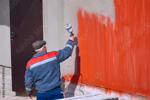 Worker paints wall facade with airless spray gun in red color. Industrial handyman painting gray wall in red. Renovating exterior of building. Pneumatic tools for spray paint work