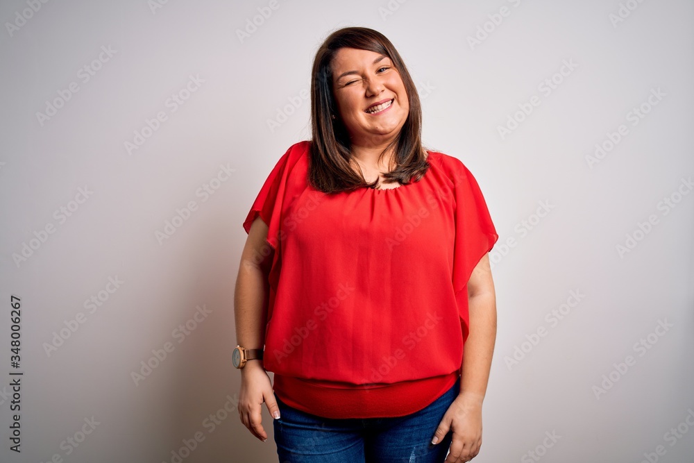 Beautiful brunette plus size woman wearing casual red t-shirt over isolated white background winking looking at the camera with sexy expression, cheerful and happy face. Beautiful brunette plus size woman wearing casual red t-shirt over isolated white background winking looking at the camera with sexy expression, cheerful and happy face.