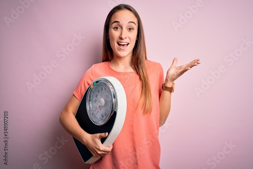 Young beautiful woman controlling weight holding weighting machine over pink background very happy and excited  winner expression celebrating victory screaming with big smile and raised hands