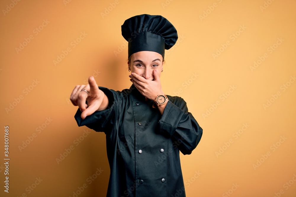 Young beautiful chef woman wearing cooker uniform and hat standing over yellow background laughing at you, pointing finger to the camera with hand over mouth, shame expression Young beautiful chef woman wearing cooker uniform and hat standing over yellow background laughing at you, pointing finger to the camera with hand over mouth, shame expression