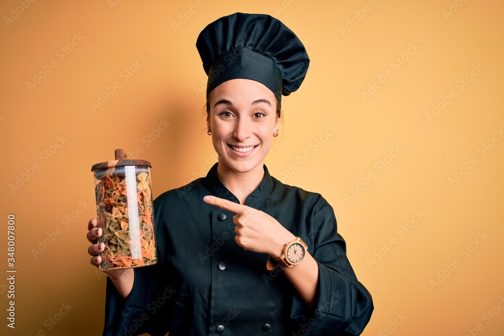 Young beautiful chef woman wearing cooker uniform and hat holding bottle with italian pasta very happy pointing with hand and finger
