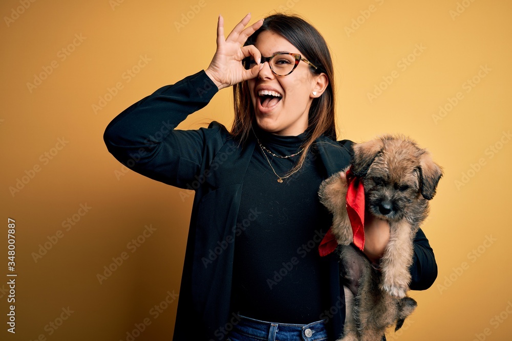 Young beautiful brunette woman holding cute puppy pet over isolated yellow background with happy face smiling doing ok sign with hand on eye looking through fingers