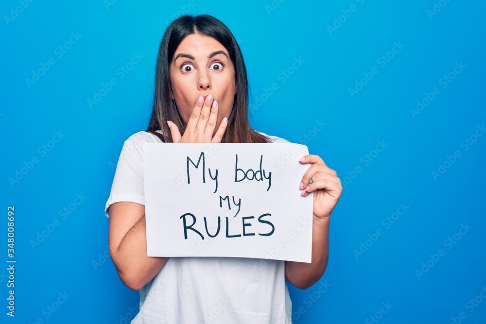 Young beautiful woman asking for women rights holding paper with my body my rules message covering mouth with hand, shocked and afraid for mistake. Surprised expression Young beautiful woman asking for women rights holding paper with my body my rules message covering mouth with hand, shocked and afraid for mistake. Surprised expression