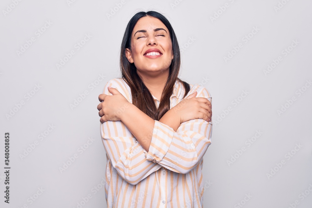 Young beautiful brunette woman wearing casual striped shirt standing over white background hugging oneself happy and positive, smiling confident. Self love and self care Young beautiful brunette woman wearing casual striped shirt standing over white background hugging oneself happy and positive, smiling confident. Self love and self care