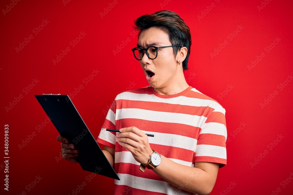 Young handsome chinese inspector man wearing glasses holding clipboard checklist scared in shock with a surprise face, afraid and excited with fear expression Young handsome chinese inspector man wearing glasses holding clipboard checklist scared in shock with a surprise face, afraid and excited with fear expression