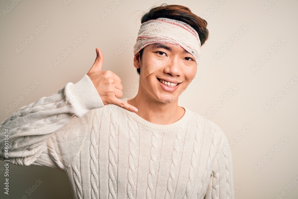 Young handsome chinese man injured for accident wearing bandage and strips on head smiling doing phone gesture with hand and fingers like talking on the telephone. Communicating concepts. Young handsome chinese man injured for accident wearing bandage and strips on head smiling doing phone gesture with hand and fingers like talking on the telephone. Communicating concepts.
