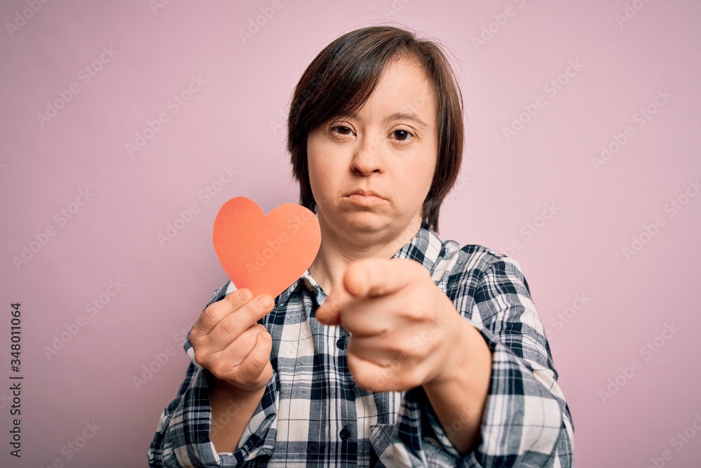 Young down syndrome romantic woman holding red heart paper shape over pink background pointing with finger to the camera and to you, hand sign, positive and confident gesture from the front Young down syndrome romantic woman holding red heart paper shape over pink background pointing with finger to the camera and to you, hand sign, positive and confident gesture from the front