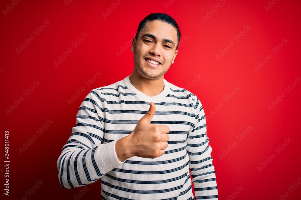 Young brazilian man wearing casual striped t-shirt standing over isolated red background doing happy thumbs up gesture with hand. Approving expression looking at the camera showing success. Young brazilian man wearing casual striped t-shirt standing over isolated red background doing happy thumbs up gesture with hand. Approving expression looking at the camera showing success.