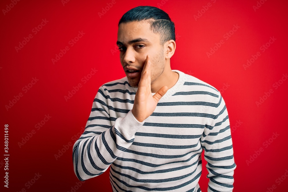 Young brazilian man wearing casual striped t-shirt standing over isolated red background hand on mouth telling secret rumor, whispering malicious talk conversation Young brazilian man wearing casual striped t-shirt standing over isolated red background hand on mouth telling secret rumor, whispering malicious talk conversation