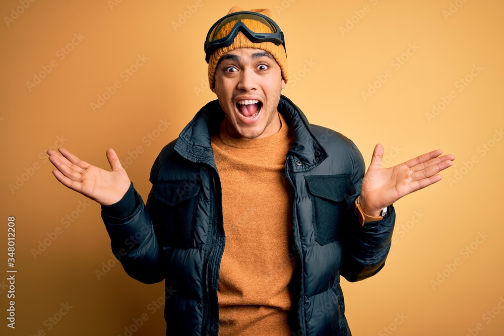 Young brazilian skier man wearing snow sportswear and ski goggles over yellow background celebrating crazy and amazed for success with arms raised and open eyes screaming excited. Winner concept Young brazilian skier man wearing snow sportswear and ski goggles over yellow background celebrating crazy and amazed for success with arms raised and open eyes screaming excited. Winner concept