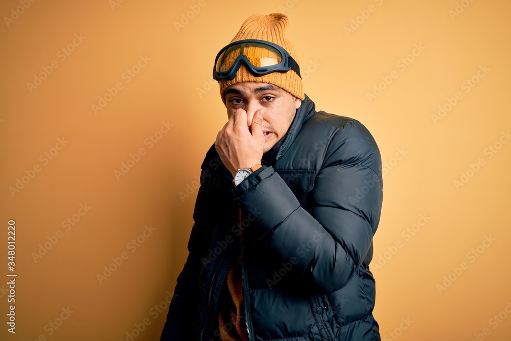 Young brazilian skier man wearing snow sportswear and ski goggles over yellow background smelling something stinky and disgusting, intolerable smell, holding breath with fingers on nose. Bad smell Young brazilian skier man wearing snow sportswear and ski goggles over yellow background smelling something stinky and disgusting, intolerable smell, holding breath with fingers on nose. Bad smell