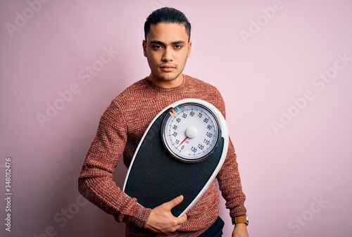Young brazilian man doing diet to lose weigth holding scale over isolated pink background with a confident expression on smart face thinking serious photo