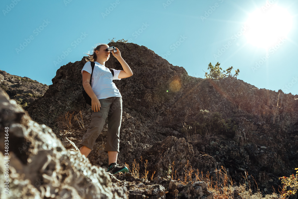 Traveler hiking girl with backpack and holding binoculars looking on top of mountain lit by sun