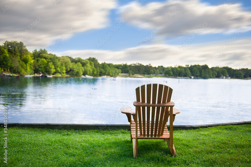Adirondack chair on a green grass carpet facing a calm lake. Across the water are cottages nestled among green trees. Adirondack chair on a green grass carpet facing a calm lake. Across the water are cottages nestled among green trees.