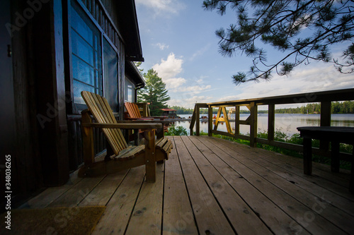 Adirondack chairs on a wooden porch facing a calm lake. In the background there are lounge chairs on a wooden dock.
