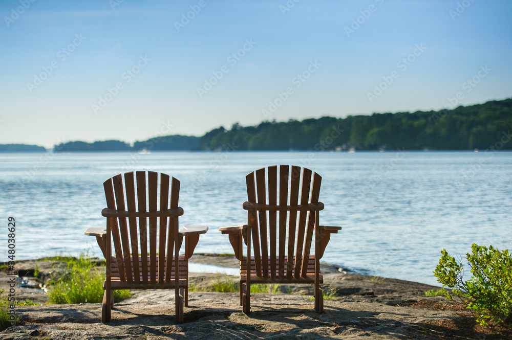 Two Adirondack chairs sit on a rock formation facing the waters of a lake during a sunny summer day in Muskoka, Ontario Canada.