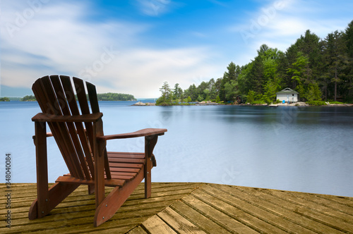 Muskoka chair sitting on a wood dock facing a lake. Across the calm water is a white cottage nestled among green trees. There is a boat dock on the water in front of the cottage.