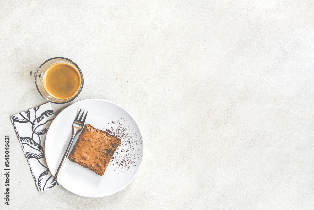 Top view of a cup of coffee and a dessert plate with brownie cake over white rustic background.