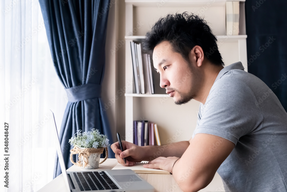 Asian businessman working from home in living room due to covid 19 pandemic. Man feeling tired and serious stress, thinking way to complete jobs, looking on computer laptop and writing note on paper. Asian businessman working from home in living room due to covid 19 pandemic. Man feeling tired and serious stress, thinking way to complete jobs, looking on computer laptop and writing note on paper.