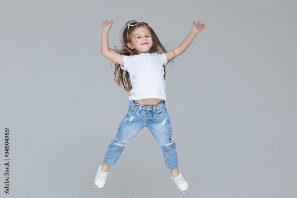Kid girl preschooler in blue jeans, white t-shirt and sunglasses is jumping with raised hands isolated on grey background Kid girl preschooler in blue jeans, white t-shirt and sunglasses is jumping with raised hands isolated on grey background
