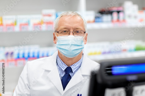 medicine, healthcare and people concept - senior apothecary wearing face protective medical mask for protection from virus disease at pharmacy cash register