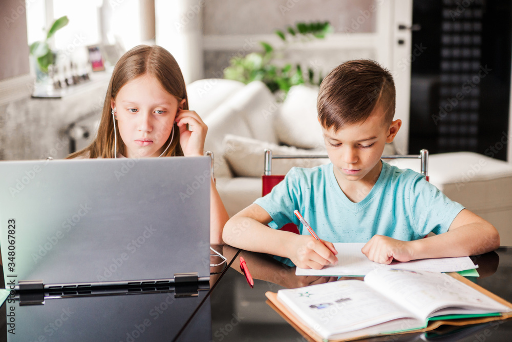 Boy and girl, brother and sister study at home. A girl watches a video lesson or an online conference, a boy does exercises in a notebook from a textbook Boy and girl, brother and sister study at home. A girl watches a video lesson or an online conference, a boy does exercises in a notebook from a textbook