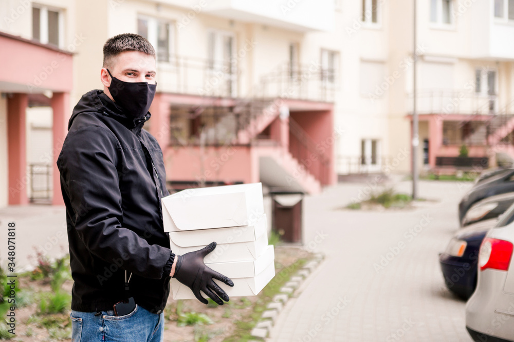 Food delivery man in black uniform, black protective mask and gloves delivers food to the house in white boxes. Horizontal photo Food delivery man in black uniform, black protective mask and gloves delivers food to the house in white boxes. Horizontal photo