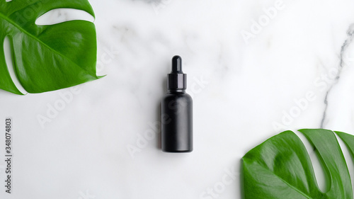 Black dropper bottle on marble background with monstera palm leaf. Blank aromatic oil container design, medical packaging template. Herbal cosmetic concept. Flat lay, top view.