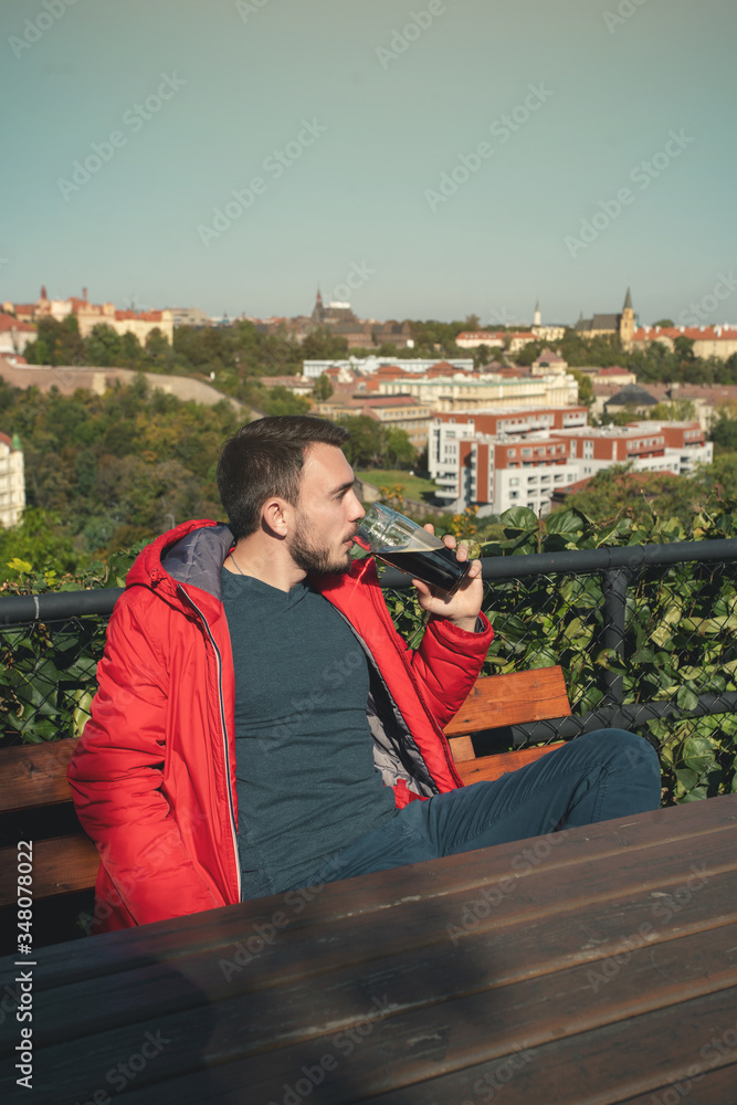 Fototapeta premium Man drinking beer. Handsome young hipster man drinking beer while sitting at the bar outdoors with panoramic view of Prague