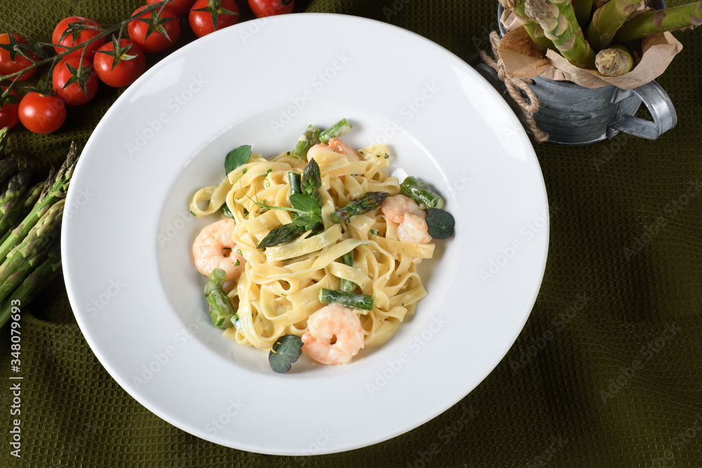 Tagliatelle or pappardelle pasta with asparagus and shrimps, cherry tomatoes in the background.
