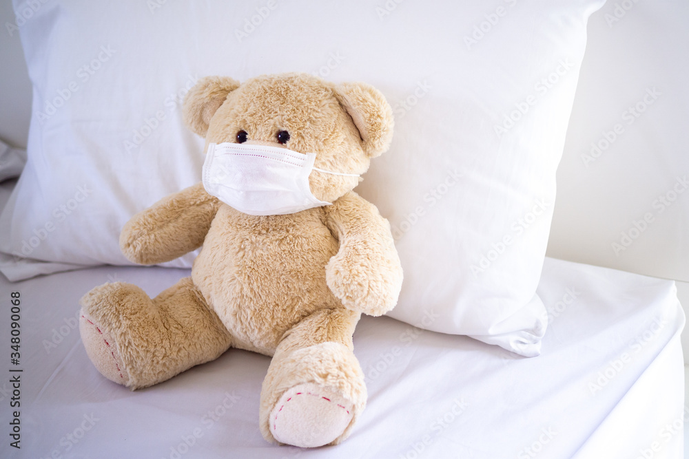 The teddy bear is sitting on a bed wearing a mask to protect against germs and viruses. Hygiene for children concept