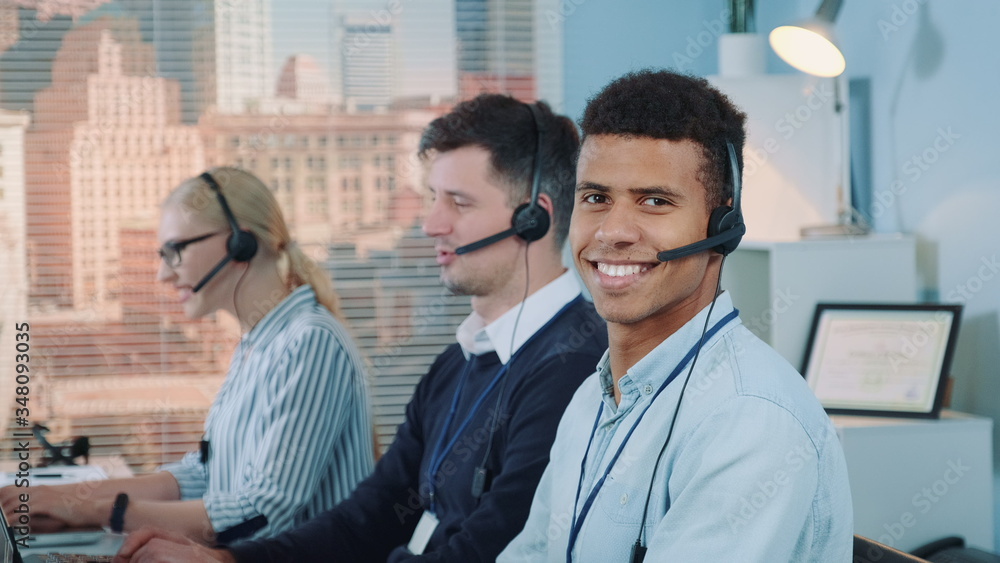 Close-up shot of smiling mixed-race man taking call in busy call center. In the background there are his colleagues also talking to clients on phone. Close-up shot of smiling mixed-race man taking call in busy call center. In the background there are his colleagues also talking to clients on phone.