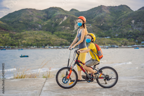 Active school kid boy and his mom in medical mask and safety helmet riding a bike with backpack on sunny day. Happy child biking on way to school. You need to go to school in a mask because of the