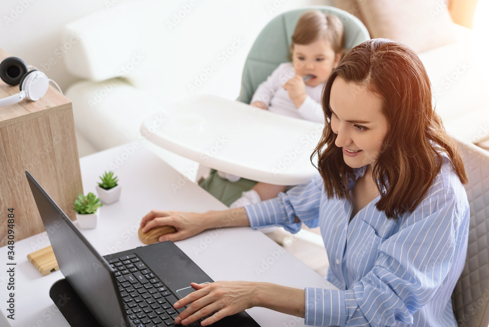 smiling woman in blue shirt is working at laptop, child sitting next to her, mother on maternity leave, freelancer, remote work at home concept smiling woman in blue shirt is working at laptop, child sitting next to her, mother on maternity leave, freelancer, remote work at home concept