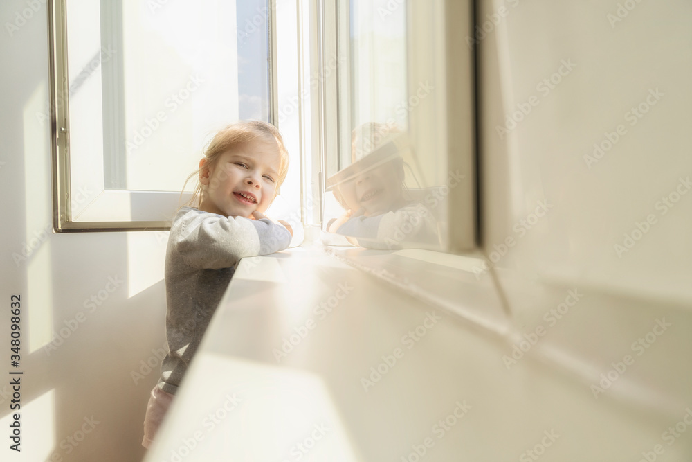 little girl dreamily looks out the window. the sun illuminates her face, flare. the child is on self-isolation and has no opportunity to walk on the street. little girl dreamily looks out the window. the sun illuminates her face, flare. the child is on self-isolation and has no opportunity to walk on the street.