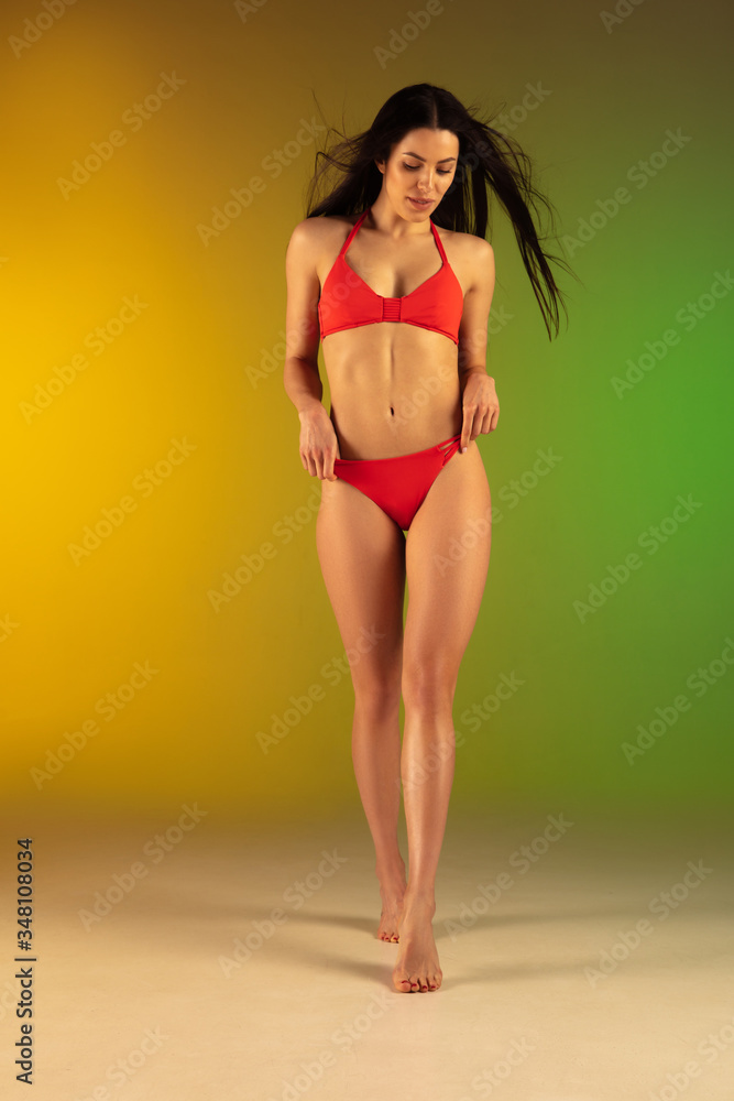 Fototapeta premium Fashion portrait of young fit and sportive caucasian woman in stylish red swimwear on gradient background. Brunette longhair model. Perfect body ready for summertime. Beauty, resort, sport concept.