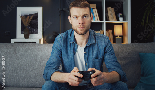 Front view of serious young man sitting on sofa playing video games using remote control joystick