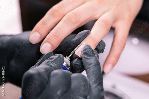 Close-up professional beautician hands working with electric drill on client fingernails. Procedure applying artificial fingernails. Hands of manicurist with tool. Manicurist in gloves removes cuticle