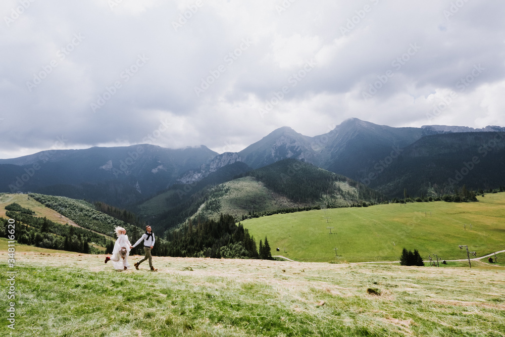 Beautiful bride in a boho style dress and groom walk on the field near the mountains. Wedding photo shoot in the mountains.